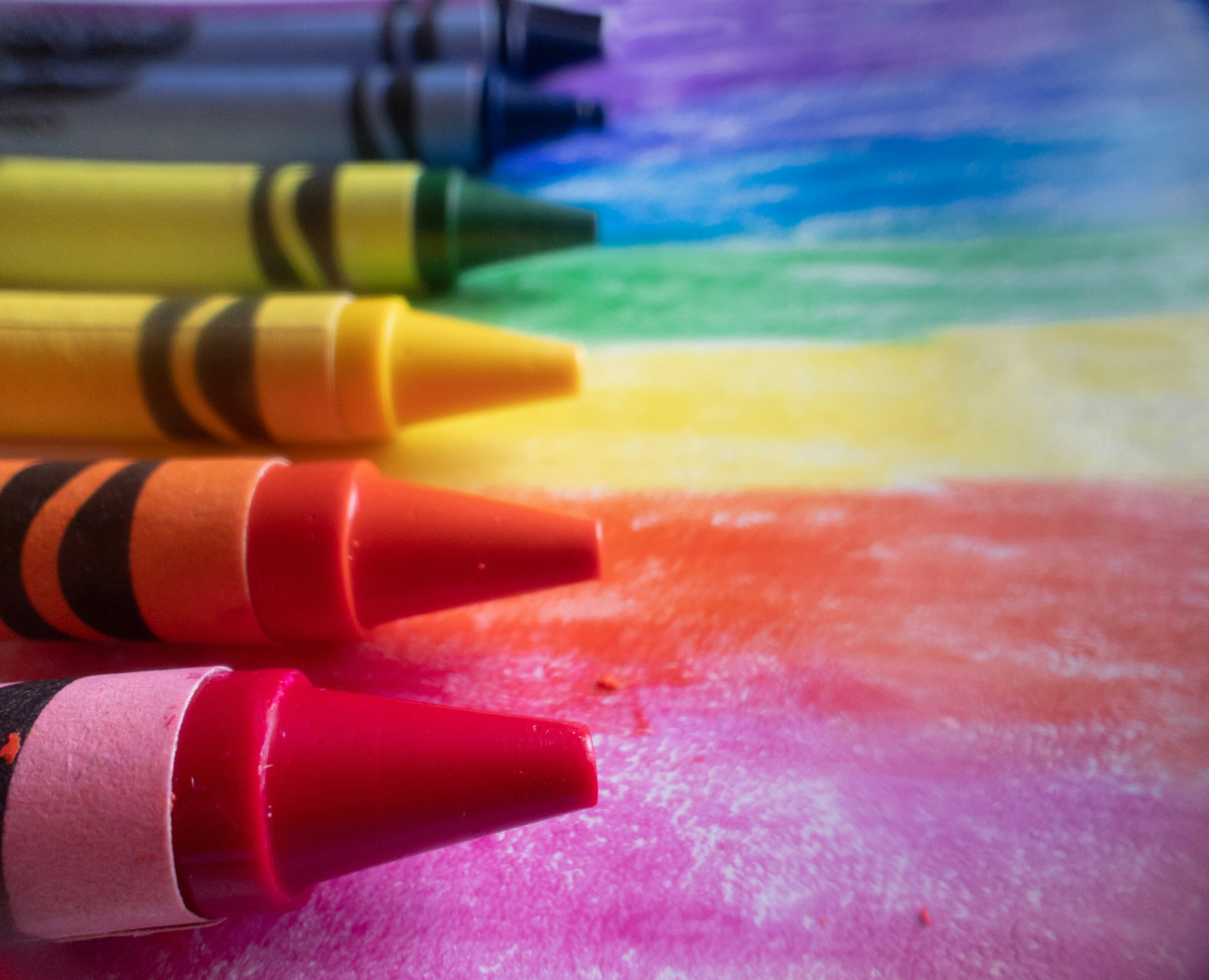 A row of crayons in vibrant colors arranged on a rainbow background, showcasing their tips against a colorful surface.