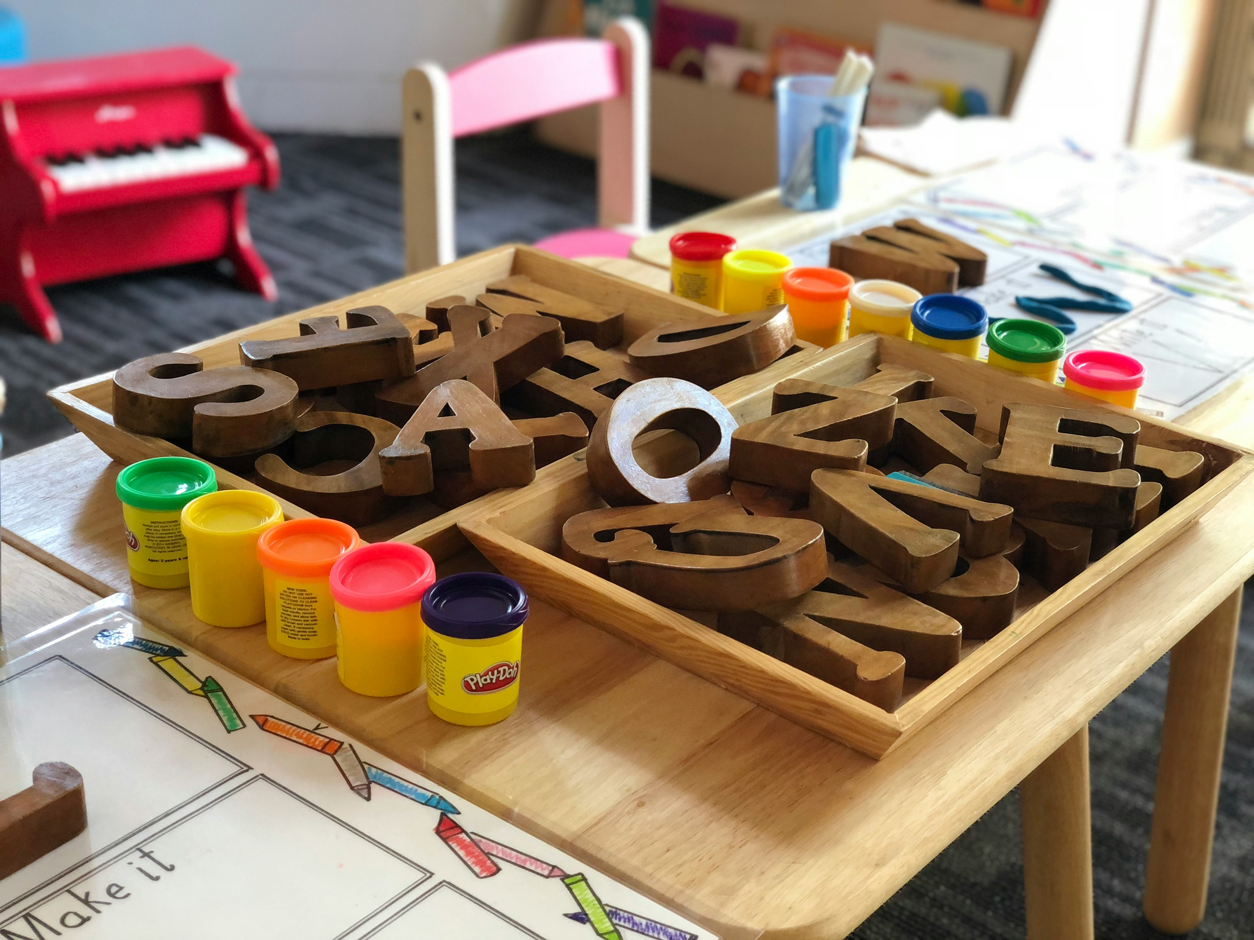 A wooden tray holds various letters and colorful Play-Doh containers on a table in a bright learning environment.