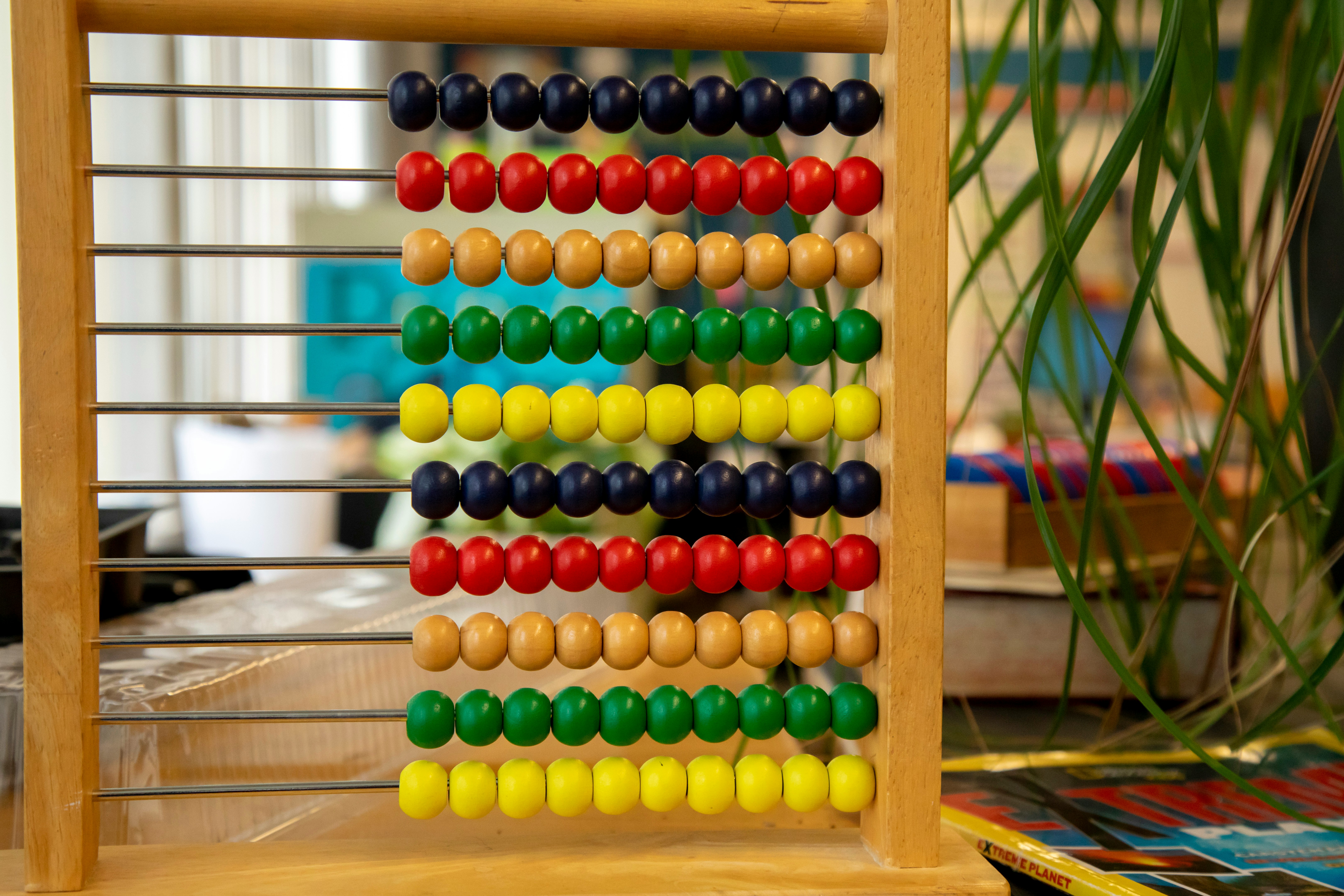 Image of a wooden abacus with ten rows, each containing ten colored beads in red, yellow, green, and blue, placed on a table with greenery in the background.